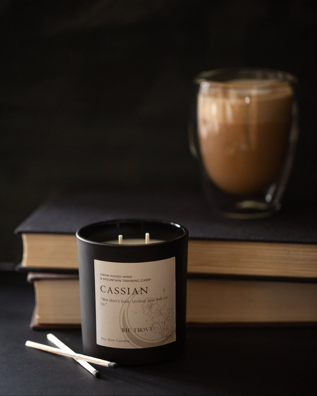 Candle labeled 'Cassian' with a cup of coffee and books in the background on a dark surface.