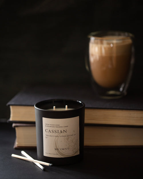 Candle labeled 'Cassian' with a cup of coffee and books in the background on a dark surface.