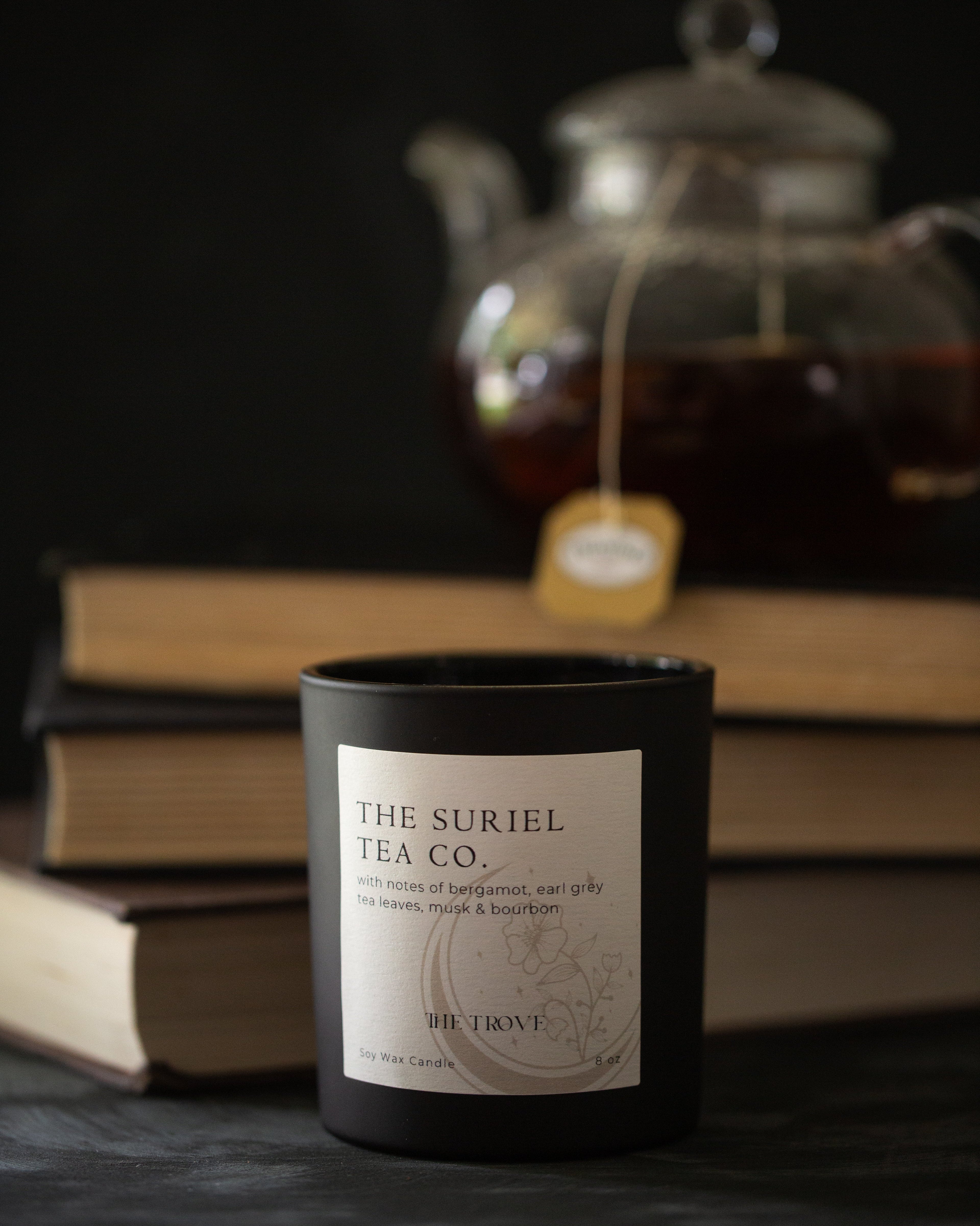 candle in black jar with 'The Suriel Tea Co.' label in front of a teapot and books on a dark background