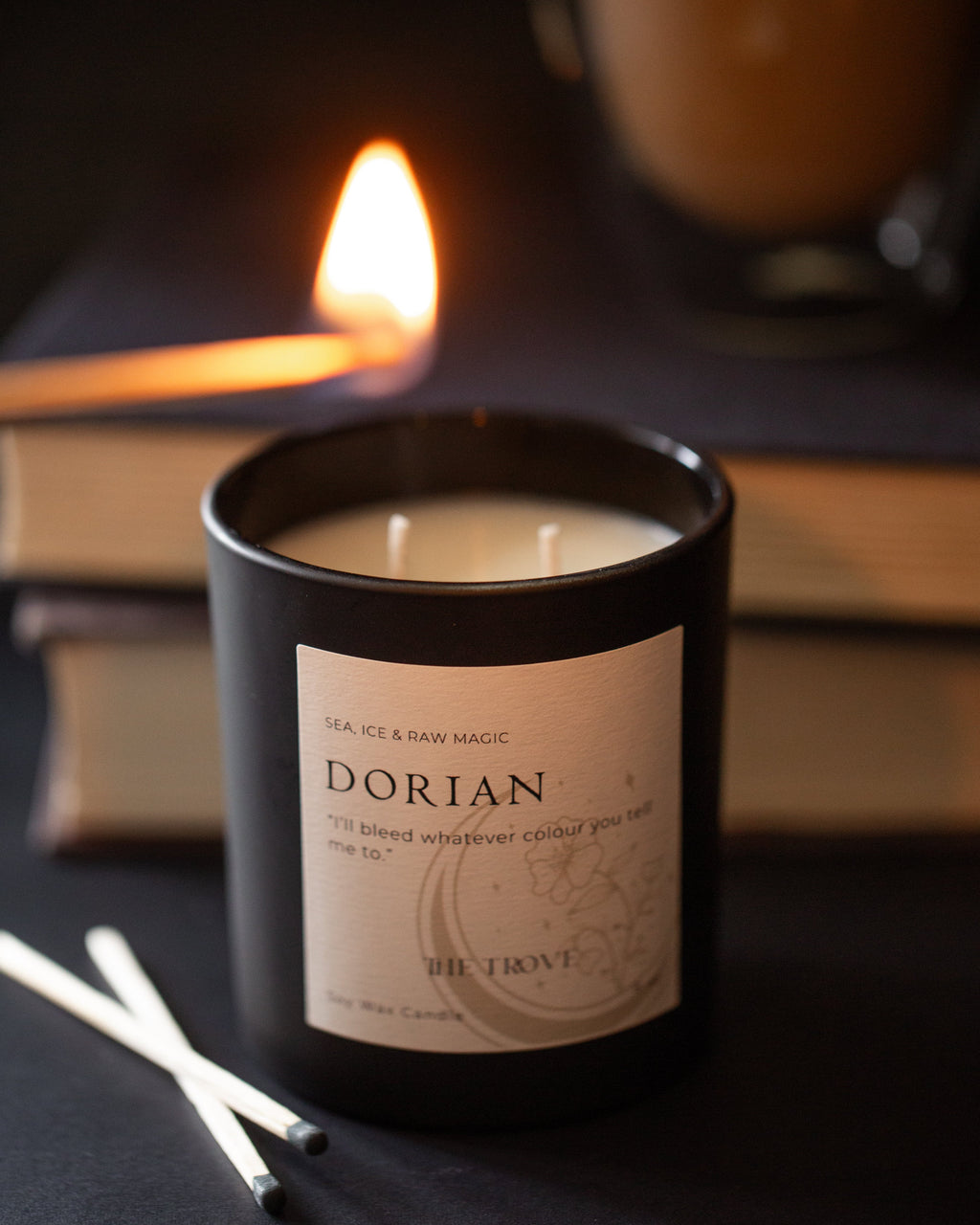 Candle in a black container labeled 'Dorian' with a lit candle and books in the background.