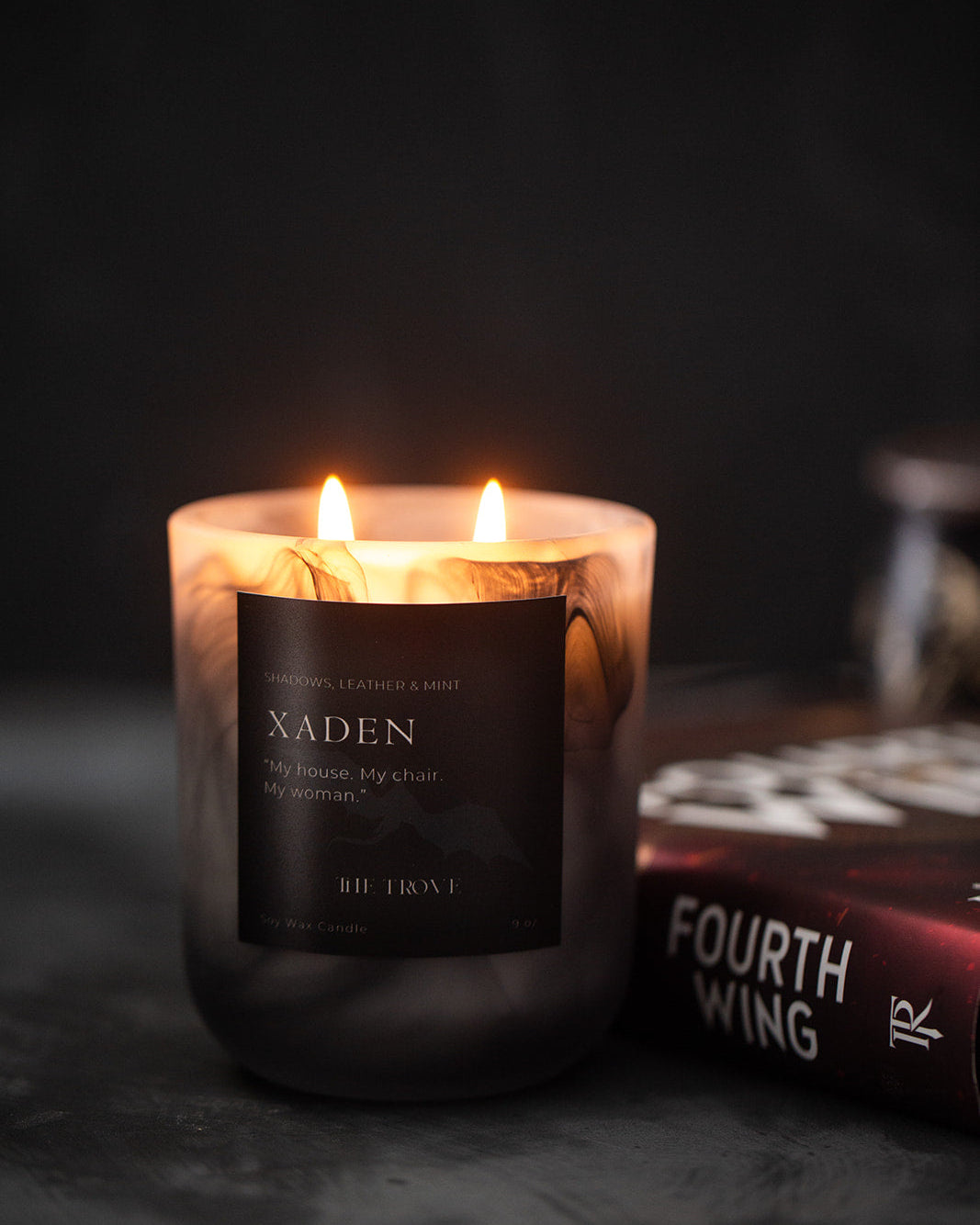 Candle labeled 'Xaden' with a book titled 'Fourth Wing' in the background on a dark surface.