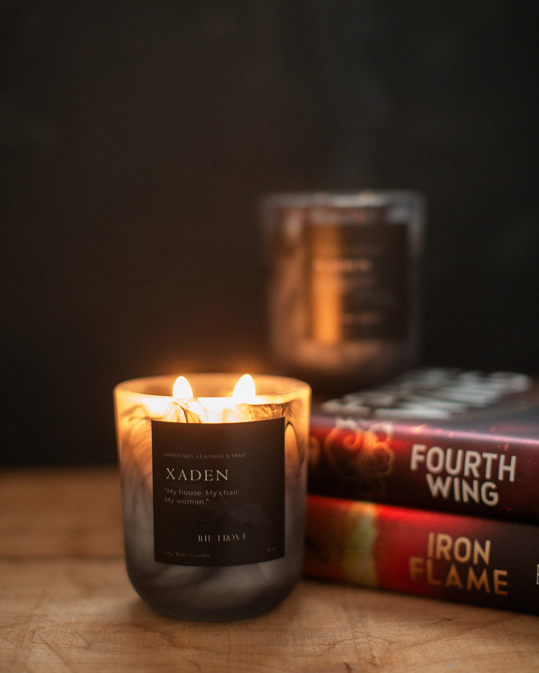 Candle labeled 'Xaden' with books in the background on a wooden surface