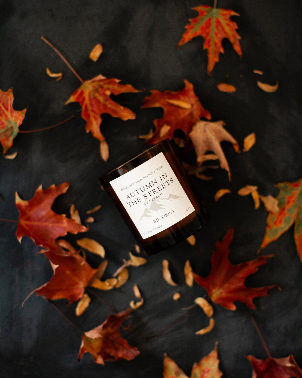 Candle labeled 'Autumn in the Streets' surrounded by autumn leaves on a dark surface