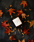 Candle labeled 'Autumn in the Streets' surrounded by autumn leaves on a dark surface