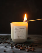 Candle labeled 'The Literary Brew' being lit with coffee beans around it on a dark background