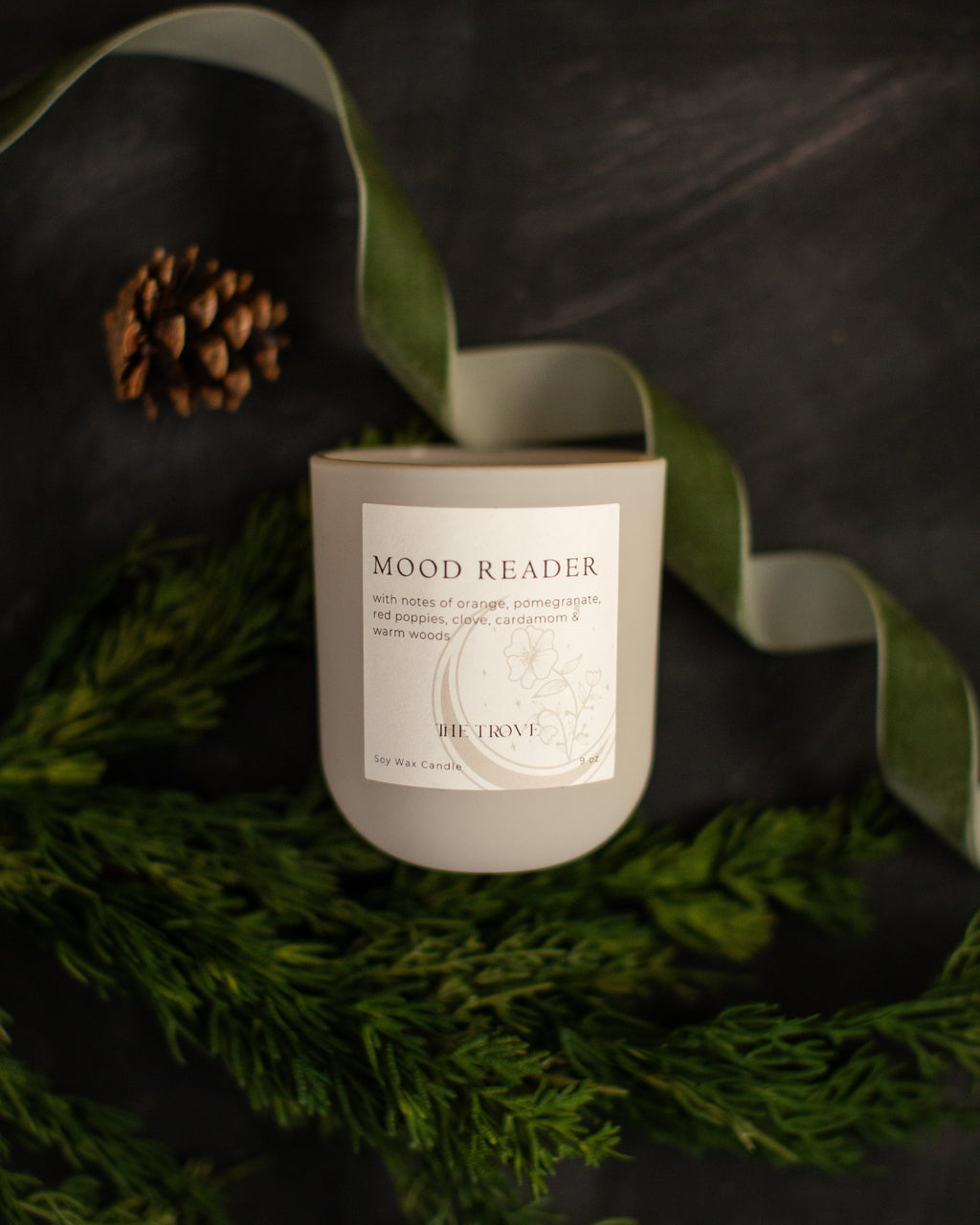Candle labeled 'Mood Reader' on a dark surface with greenery and pinecones