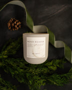 Candle labeled 'Mood Reader' on a dark surface with greenery and pinecones