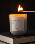 Candle labeled 'Slow Burn' being lit with a match on a dark background