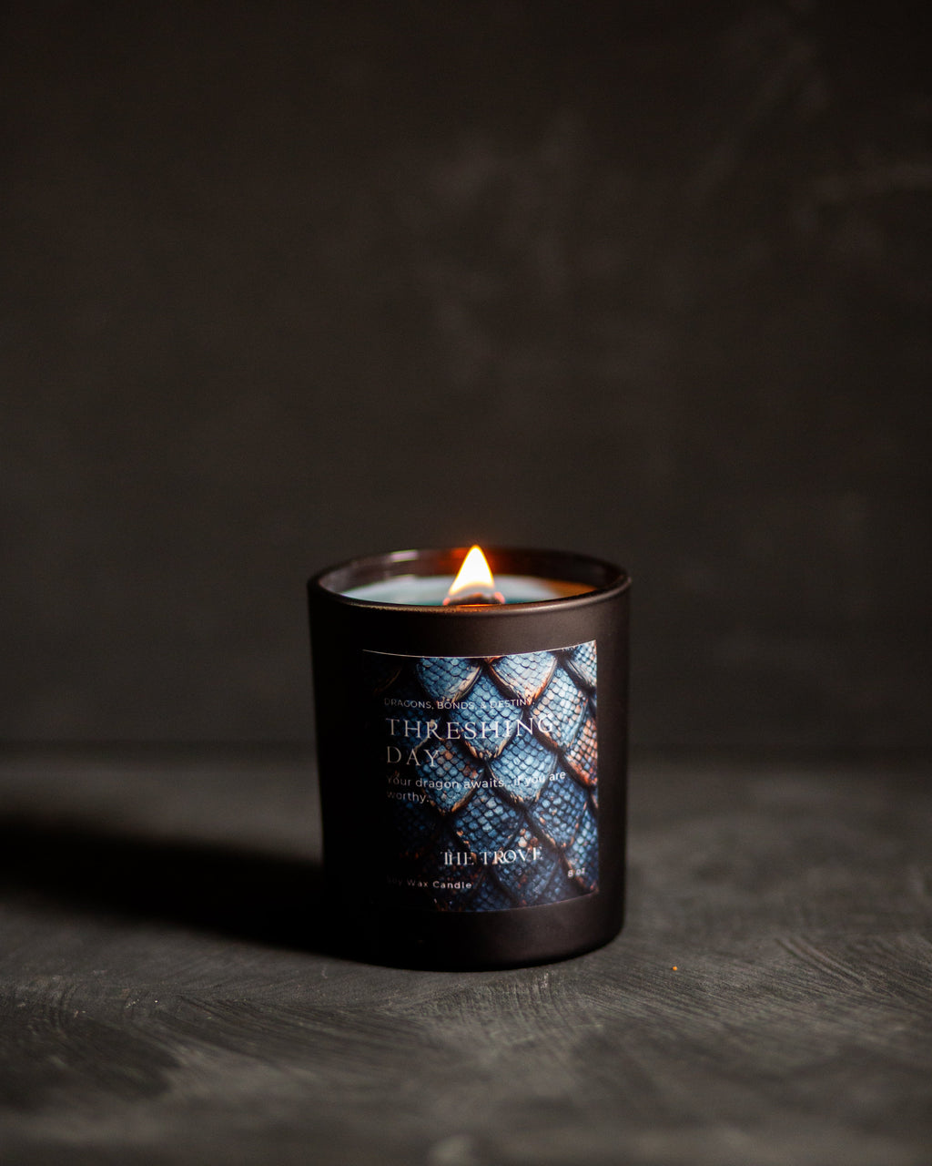 A lit candle in a dark glass jar with a blue and black patterned dragon scales label reading 'Threshing Day' set on a dark textured surface against a dark background.