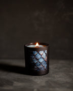 A lit candle in a dark glass jar with a blue and black patterned dragon scales label reading 'Threshing Day' set on a dark textured surface against a dark background.