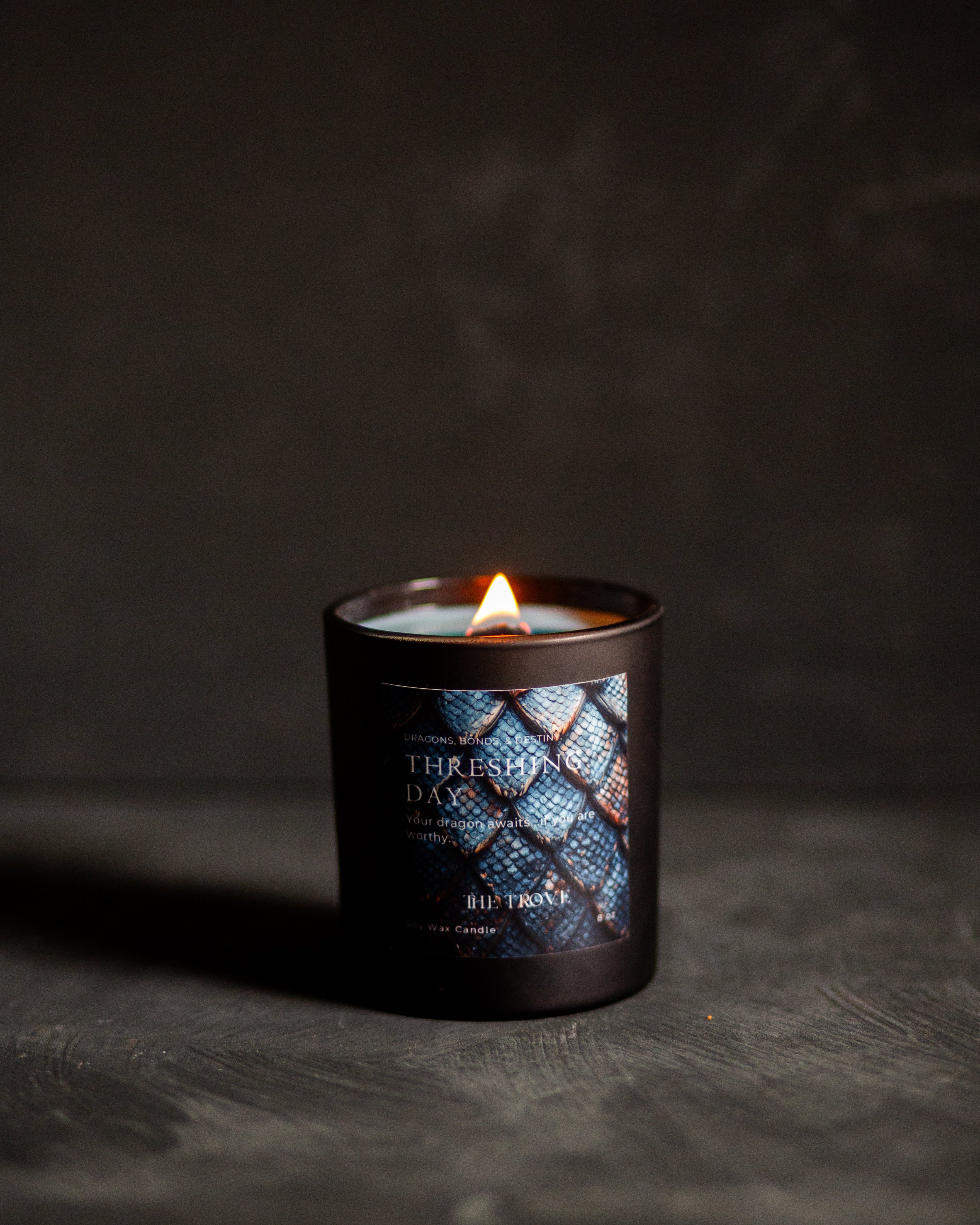 A lit candle in a dark glass jar with a blue and black patterned dragon scales label reading 'Threshing Day' set on a dark textured surface against a dark background.