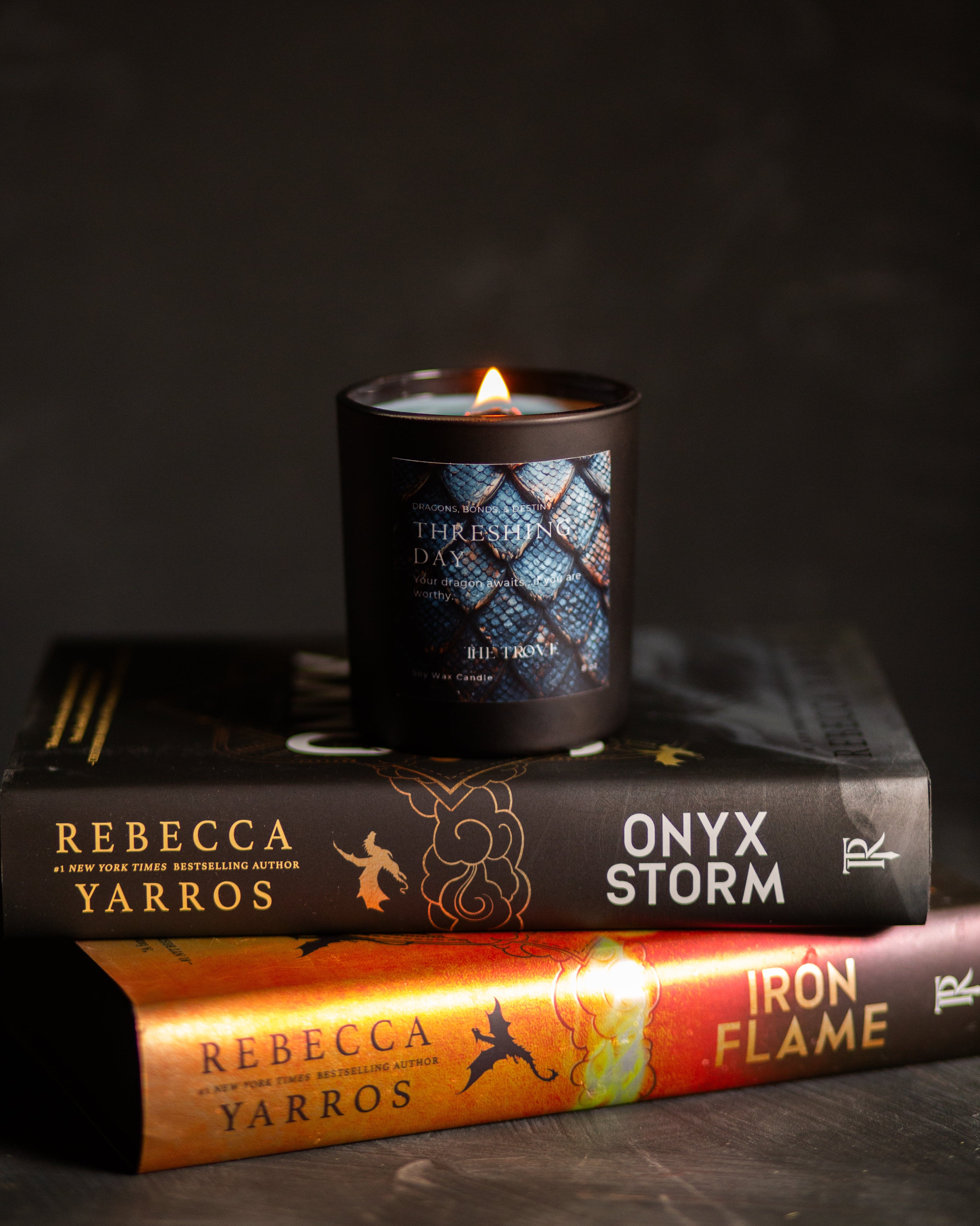 A lit black candle jar with a blue patterned label sits on top of two books, one black and one orange, against a dark background.
