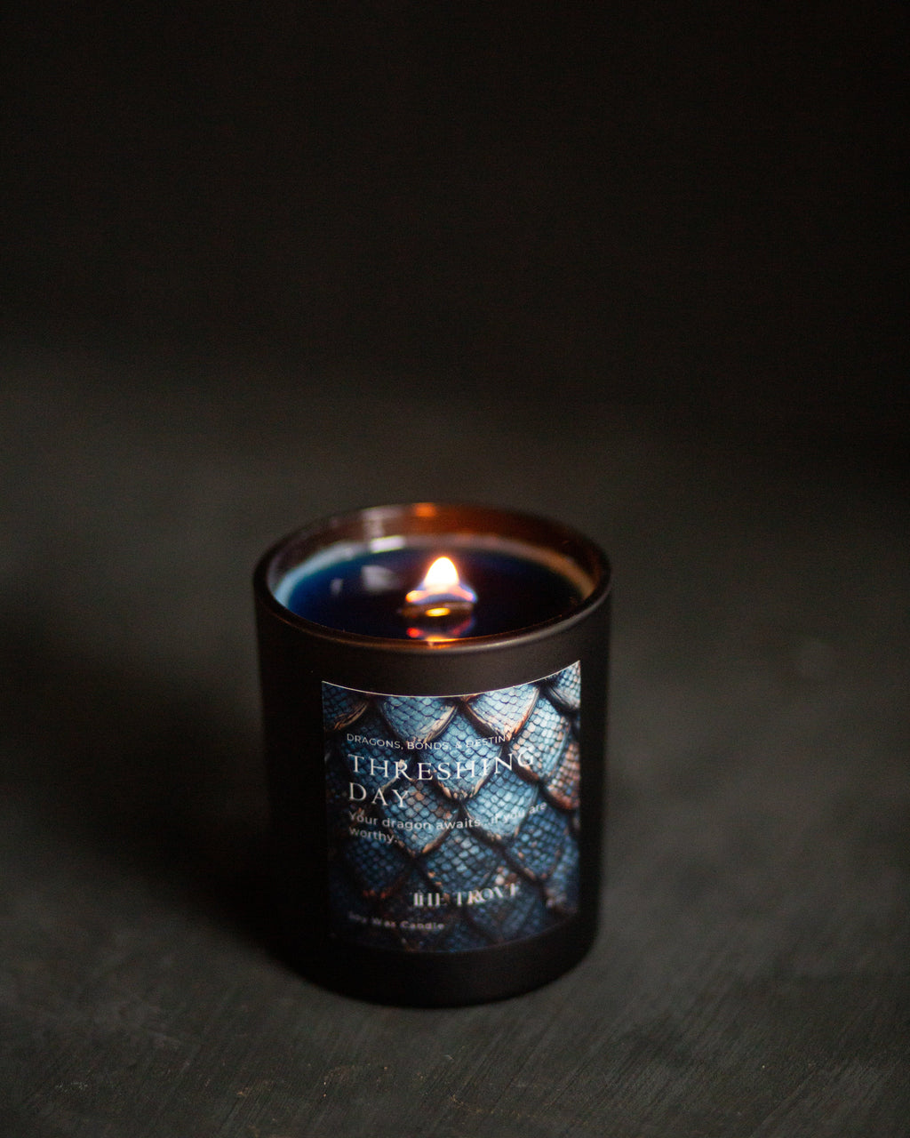 A lit candle in a dark amber glass jar with a black base, featuring a label with blue dragon scales and the text "threshing Day)