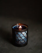 A lit candle in a dark amber glass jar with a black base, featuring a label with blue dragon scales and the text "threshing Day)