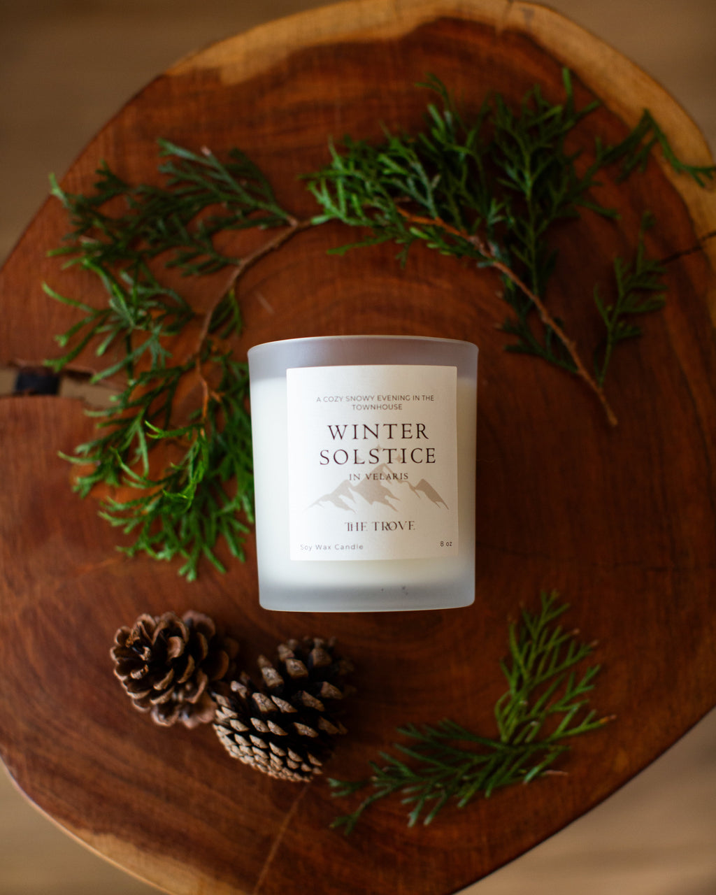 Candle labeled 'Winter Solstice' on a wooden surface with greenery and pinecones.