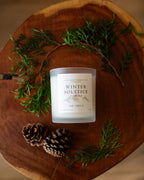 Candle labeled 'Winter Solstice' on a wooden surface with greenery and pinecones.