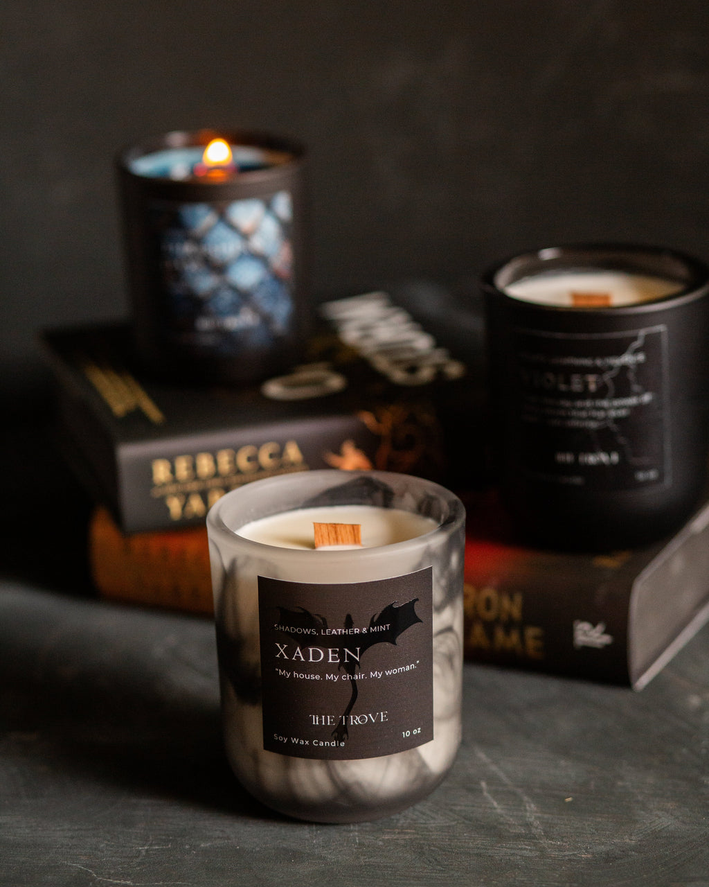 Three soy candles in dark glass jars with black labels are arranged on stacked books against a dark textured background. The front candle is lit and labeled XADEN, with two more candles behind it labeled VIOLET and another partially visible behind them.